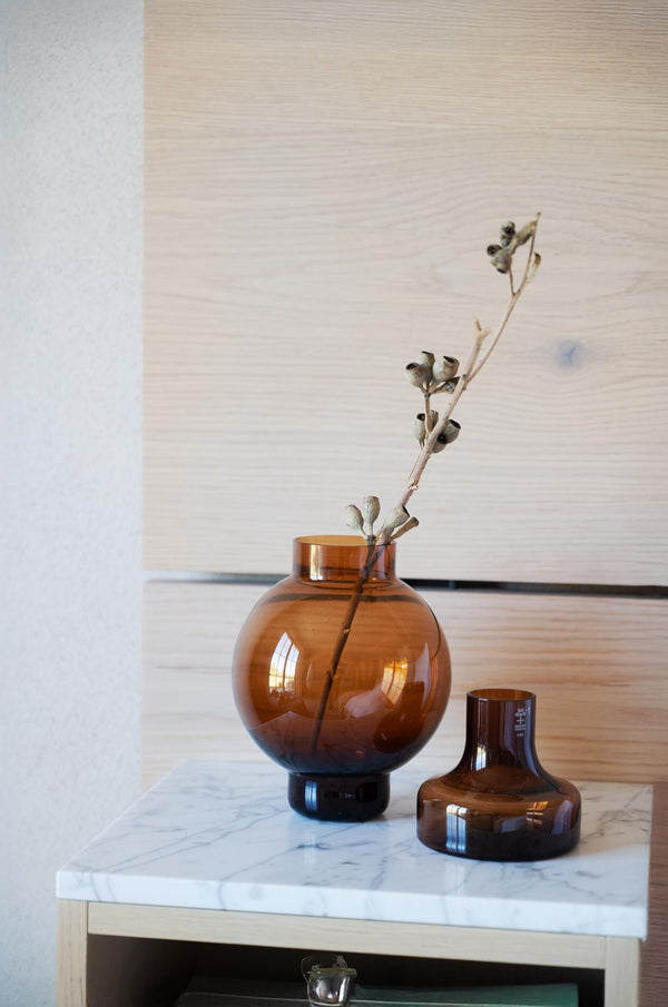 Amber glasvas med torkad eukalyptuskvist på marmorbord, bredvid en mindre brun glasvas i modern skandinavisk stil