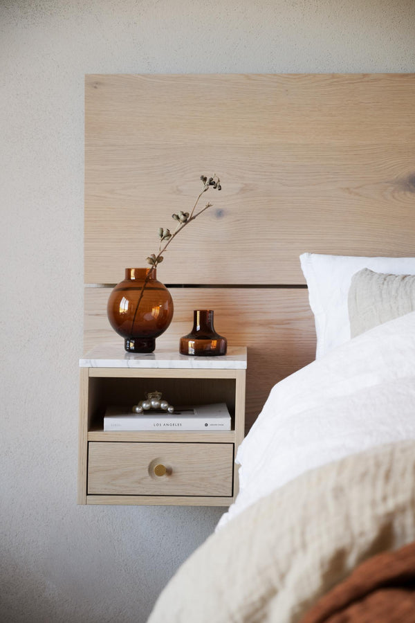 Sängbord i ljust trä med låda och öppen hylla, dekorerat med amber glasvaser och bok bredvid säng