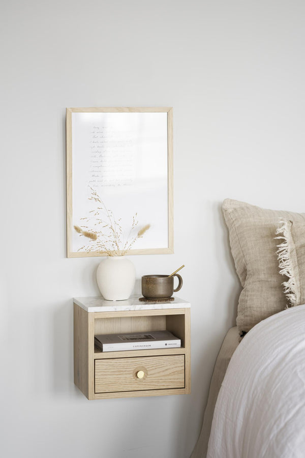 Sängbord i ljust trä med låda monterat på väggen bredvid säng med tavla och dekoration