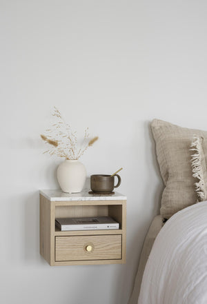 Vägghängd sängbord i ljust trä med låda och öppen hylla bredvid säng med linnekudde