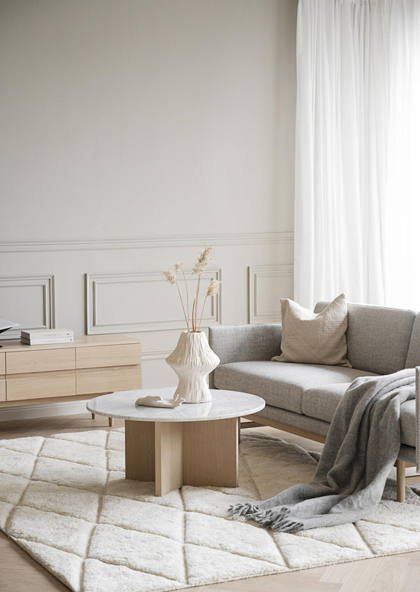 Minimalistiskt vardagsrum med beige soffa, runt marmorbord, vit vas med torkade blommor och geometrisk matta
