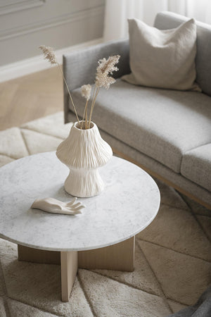 Vit texturerad vas med torkade blommor på runt marmorbord i vardagsrum med grå soffa