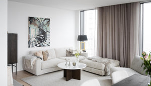 Ljust vardagsrum med beige hörnsofa, runt soffbord, grå gardiner och modern konst på väggen