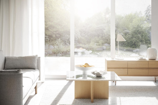 Ljust vardagsrum med grå soffa, soffbord i trä och glas, samt skänk mot fönster med trädgårdsutsikt