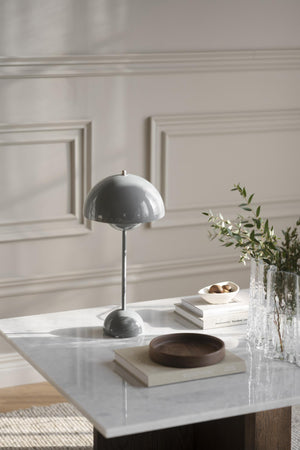 Grå bordslampa med rund skärm på vitt soffbord med böcker, skål och vas med gröna kvistar