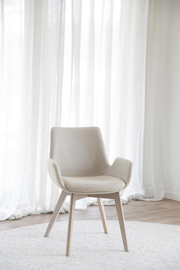 Beige stol med armstöd och träben framför vita gardiner i minimalistisk inredning