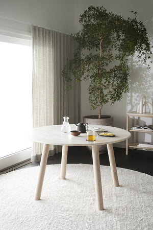 Runt matbord i ljust trä med fyra ben, placerat på ljus matta i minimalistisk matsal med grön växt