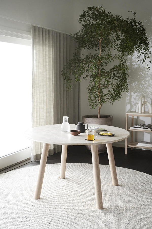 Runt matbord i ljust trä med fyra ben, placerat på ljus matta i minimalistisk matsal med grön växt