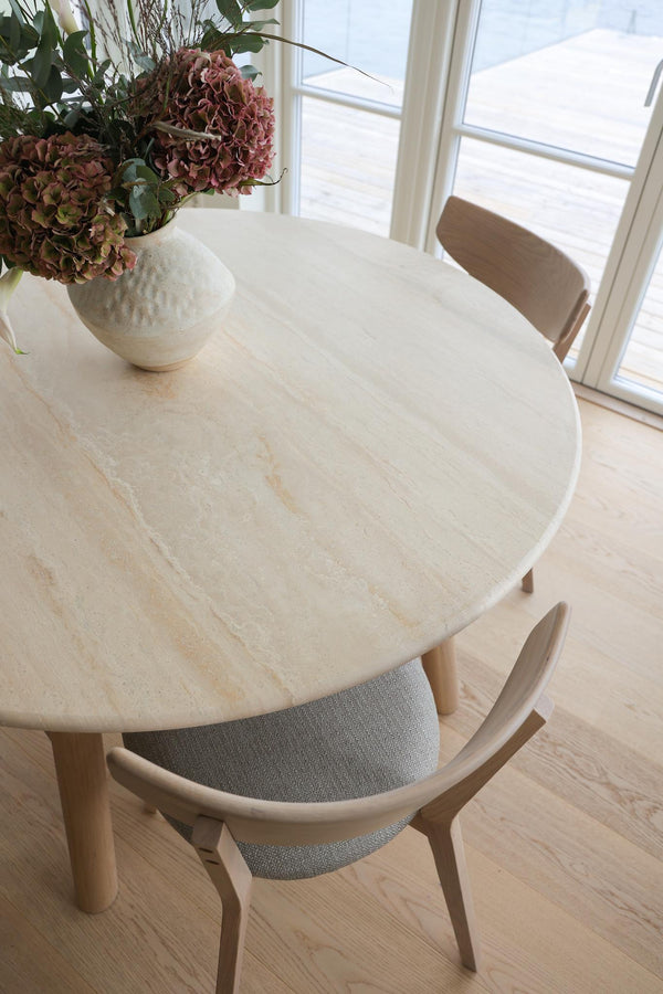 Runt matbord i ljus sten med trässtolar och vit vas med torkade blommor vid fönster