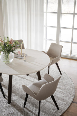 Rund matbord i ljust trä med beige stolar och svarta ben, placerat på rund matta vid fönster med vita gardiner