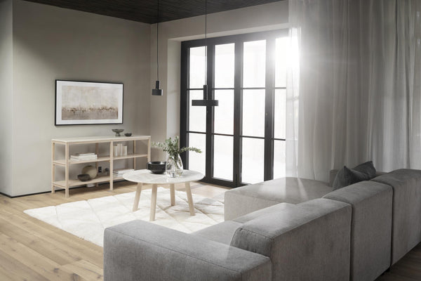 Minimalistiskt vardagsrum med grå soffa, ljust trägolv, svarta fönsterdörrar och neutral färgpalett