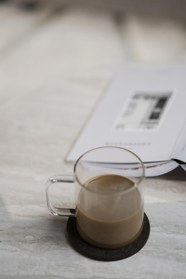 Glasmugg med handtag och guld-beige gradient, placerad på mörk underlägg med tidning i bakgrunden