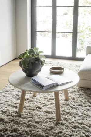 Runt soffbord i ljust trä med bok, växt och skål på fluffig matta framför fönster