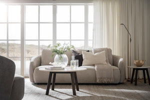 Beige tvåsitsig soffa med ljusa kuddar och pläd framför stora fönster i minimalistiskt vardagsrum