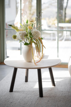 Vit vas med konstgjorda blommor på runt bord framför fönster i ljust rum