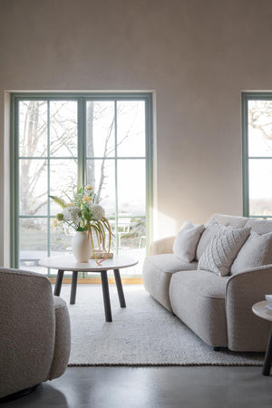 Ljust vardagsrum med grå soffa, rund soffbord och stora fönster med utsikt mot trädgård