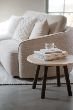Runt soffbord i ljust trä med mörka ben, dekorerat med böcker och glasmugg bredvid beige soffa