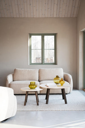 Beige boucle soffa med två runda soffbord i vitt och mörkt trä, gula glasvaser som dekoration