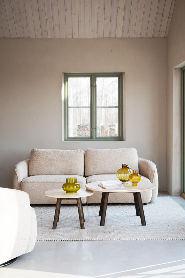 Beige boucle soffa med två runda soffbord i vitt och mörkt trä, gula glasvaser som dekoration