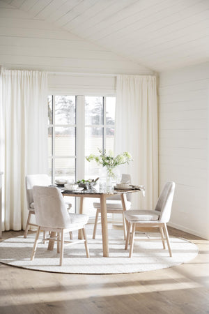 Ljus matplats med runt matbord i trä, beiga stolar och vita gardiner framför fönster med shiplap-väggar