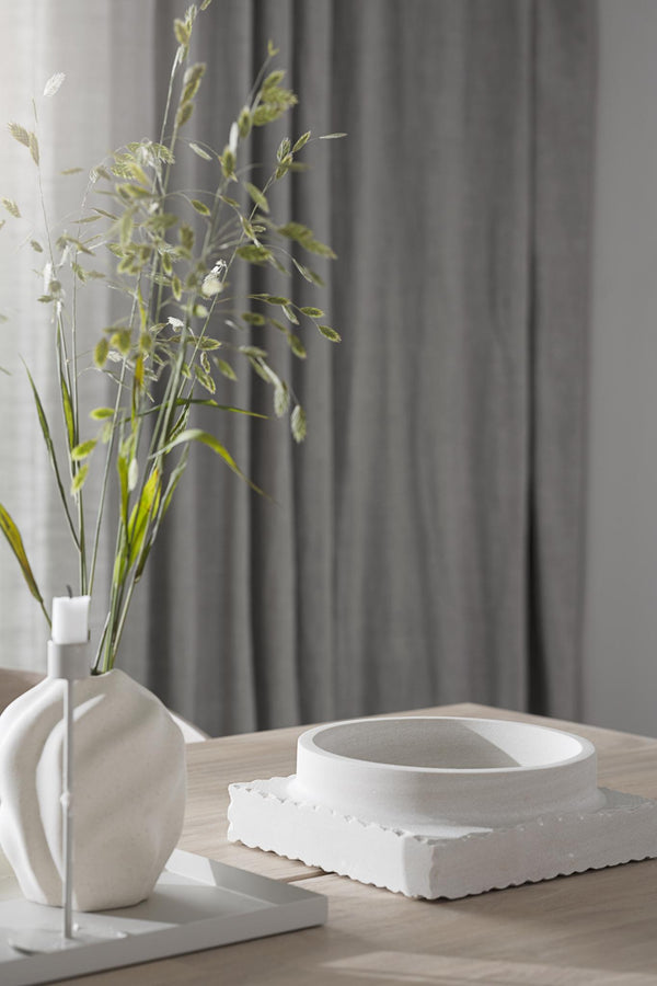 Minimalistisk stilleben med vit vas, gröna torkade blommor och keramikskål på träbord framför grå gardin