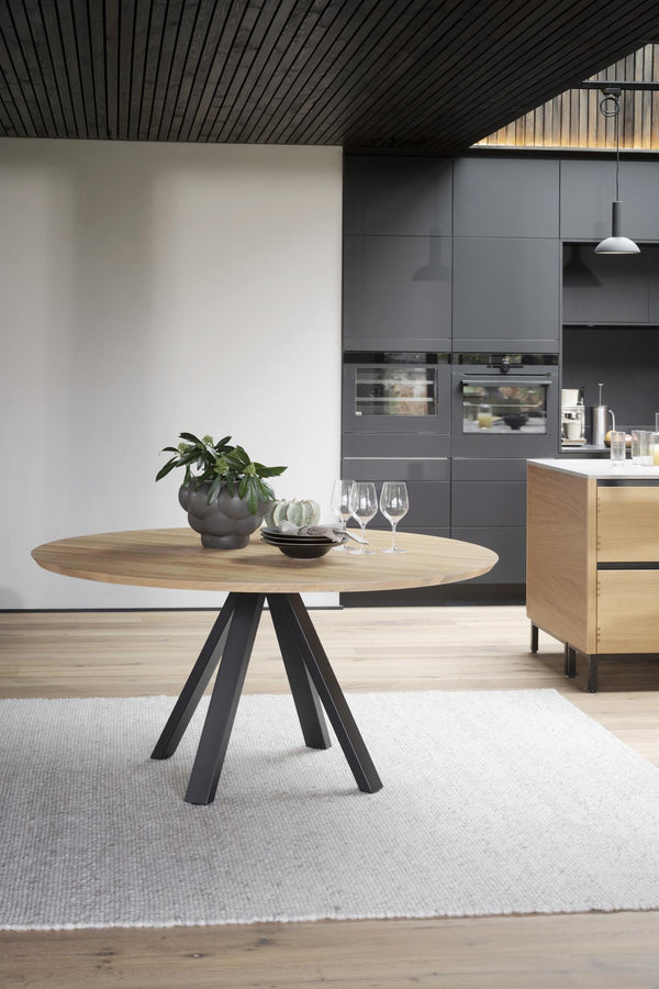 Runt matbord i trä med svarta metallben i modern matsal med grått kök i bakgrunden