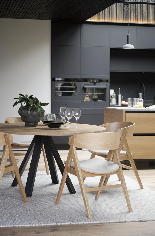 Runt matbord i ljust trä med svarta ben omgivet av moderna trässtolar i öppet kök med grå skåp
