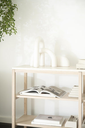 Ljust trähylla med vita böcker, tidskrifter och dekorativa skulpturer mot vit vägg med växt