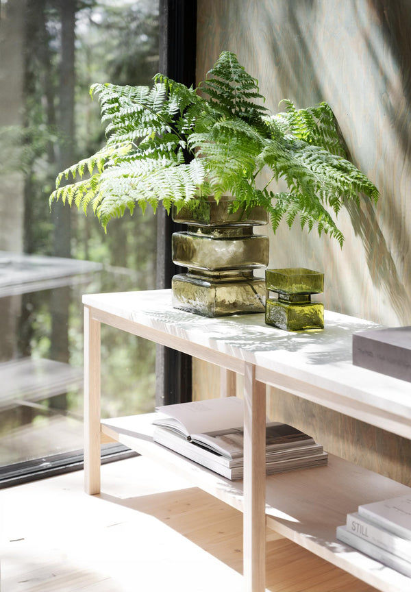 Grön glasvas med ormbunke på vit träbänk framför fönster med utsikt över skog