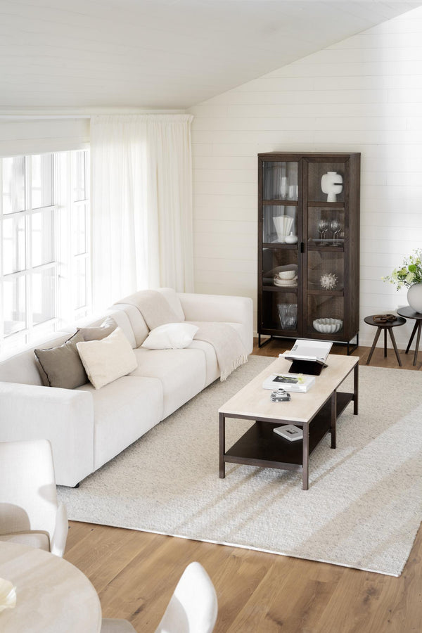 Ljust vardagsrum med vit hörnsoffa, soffbord i trä och vitrinskåp med glasdörrar