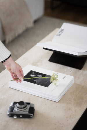 Hand som placerar en torkad blomma på en vit fotobok eller katalog på ett ljust bord med vintage kamera