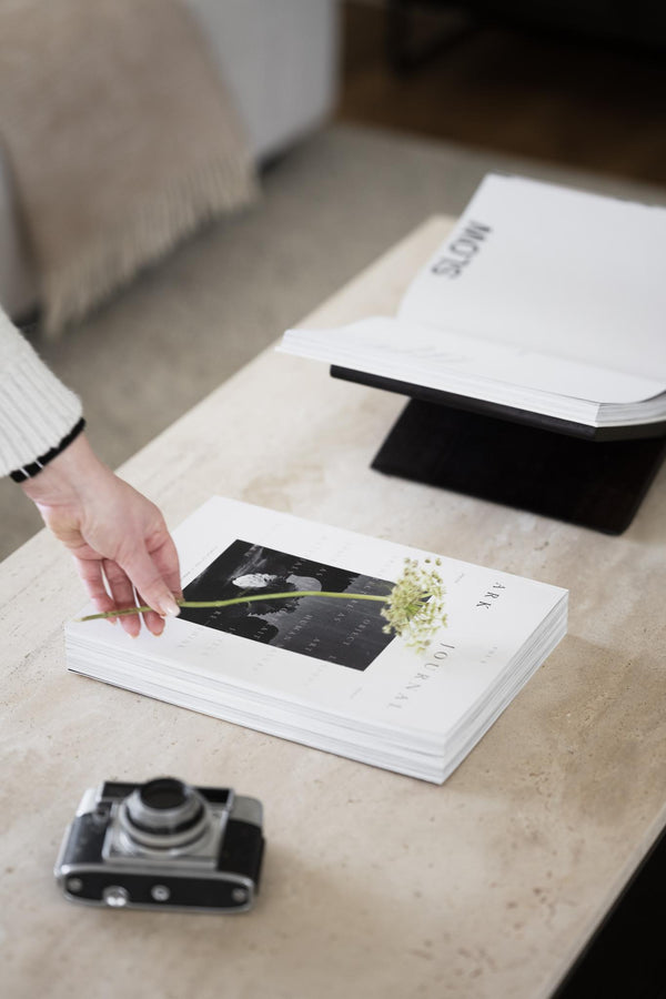 Hand som placerar en torkad blomma på en vit fotobok eller katalog på ett ljust bord med vintage kamera