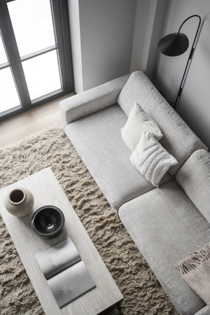 Minimalistiskt vardagsrum med grå soffa, vit soffbord, beige fluffmatta och svart golvlampa vid fönster