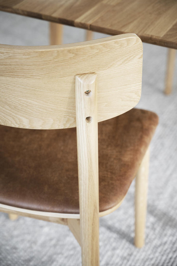 Närbild på stolsrygg i ljus ek med synliga snickardetaljer och brunt läderklädsel på sits