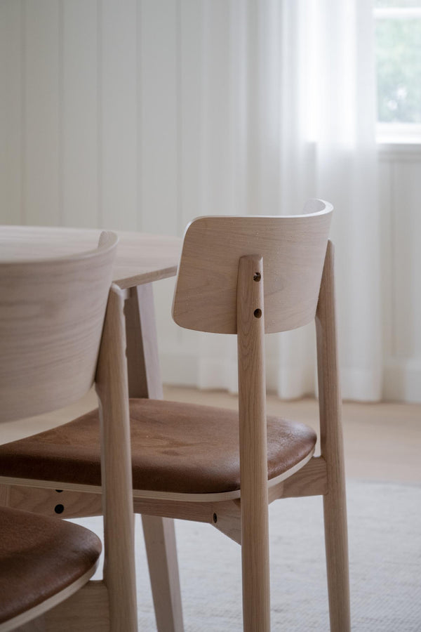 Minimalistisk matstol i ljust trä med kurvad rygg och brunt läderöverdrag vid matbord