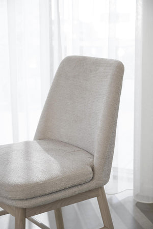 Beige stoppad stol med träben i skandinavisk design, placerad framför vit gardin i ljust rum