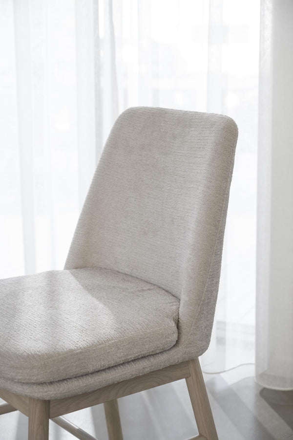 Beige stoppad stol med träben i skandinavisk design, placerad framför vit gardin i ljust rum