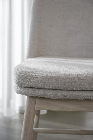 Grå stol med mjuk klädsel och träben i ljust trä vid fönster med vit gardin