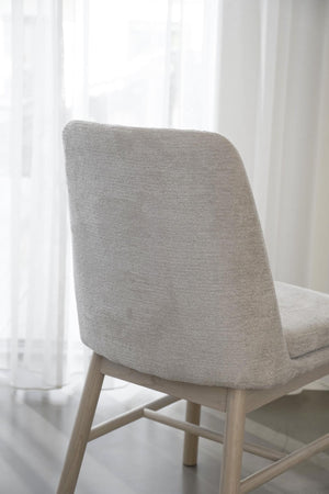 Grå stoppad stol med träben i ljust trä framför vita gardiner i minimalistisk skandinavisk stil