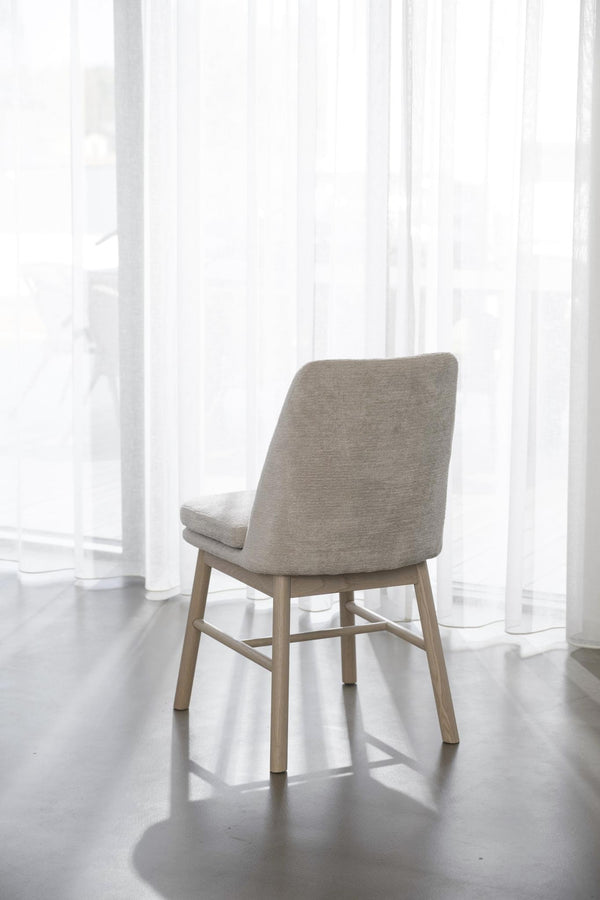 Grå stoppad stol med träben från baksidan, placerad framför vita gardindraperier i ljust rum