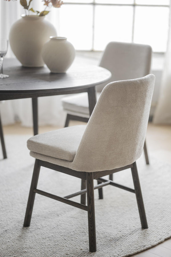 Beige matstol med mörka träben vid runt matbord med vita vaser i minimalistisk matsal