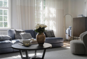 Ljust vardagsrum med blå soffa, rund soffbord, vita blommor och genomskinliga gardiner vid fönster