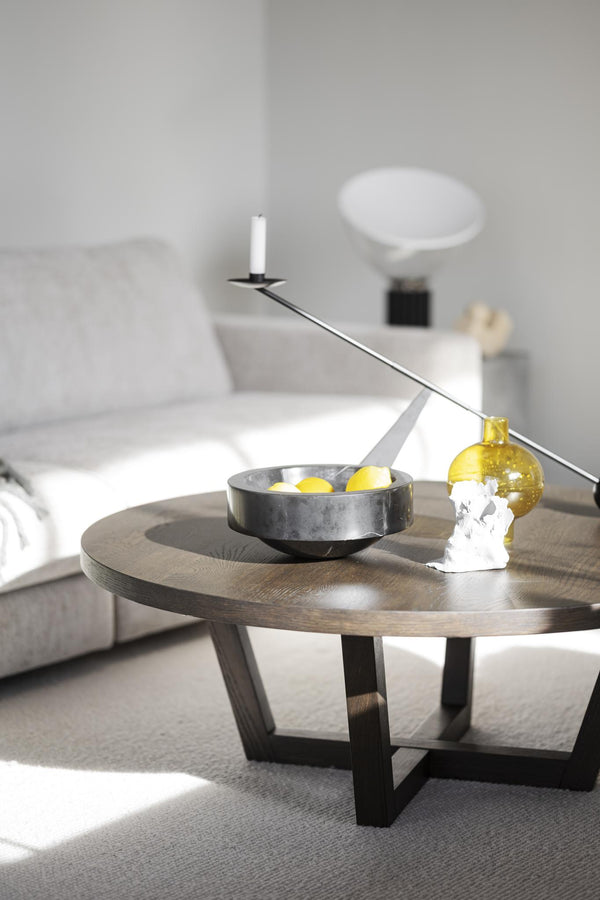 Runt soffbord i mörk ek med svart metallunderrede, dekorerat med skål, ljusstake och gul vas i minimalistiskt vardagsrum