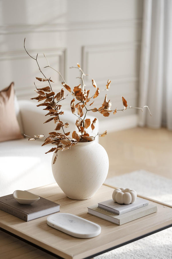 Cremefärgad keramikvas med torkade bruna löv på träbord med böcker och dekorativa skålar i ljust vardagsrum