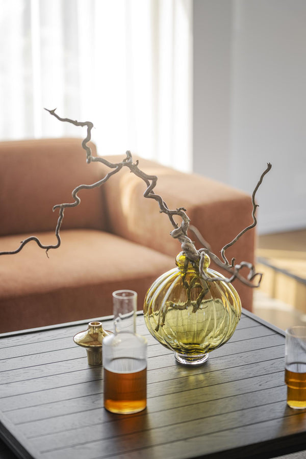 Dekorativ glasvas i gult med grenar på träbord, med flaskor i förgrunden och beige soffa i bakgrunden