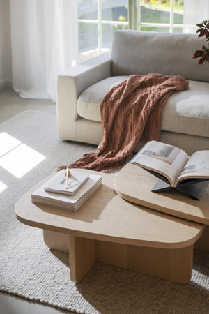Soffbord i ljust trä med organisk form, dekorerat med böcker och bredvid beige soffa med terrakottafärgad filt