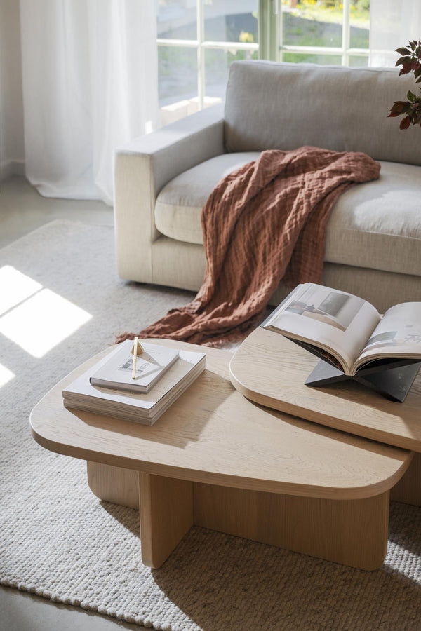 Soffbord i ljust trä med organisk form, dekorerat med böcker och bredvid beige soffa med terrakottafärgad filt