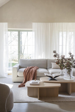 Ljust vardagsrum med beige soffa, runda träbord, vita gardiner och högt i tak