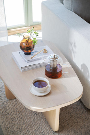 Ljust träsoffbord med tekanna, tekopp, böcker och vas med växt bredvid grå soffa