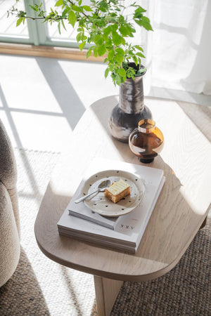 Ljust vardagsrum med träsoffbord, keramikvas med gröna växter, bärnstensfärgad glasvas och tårta på fat